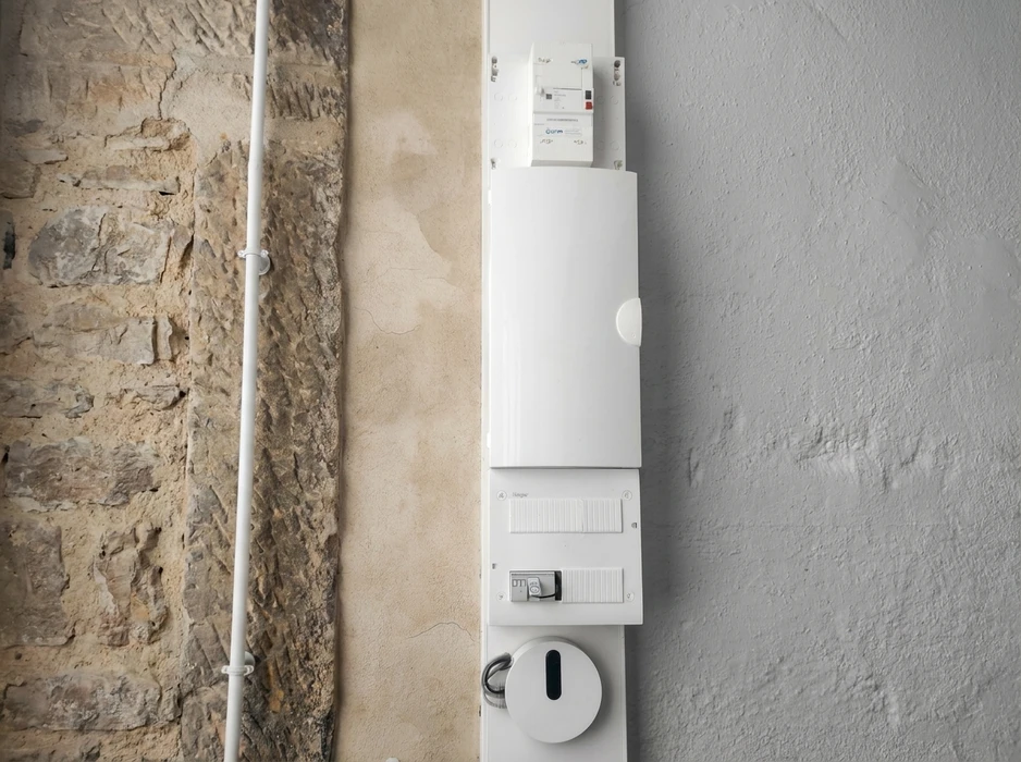 Tableau électrique résidentiel mural blanc, installé entre deux murs contrastés (pierre apparente et enduit lisse)