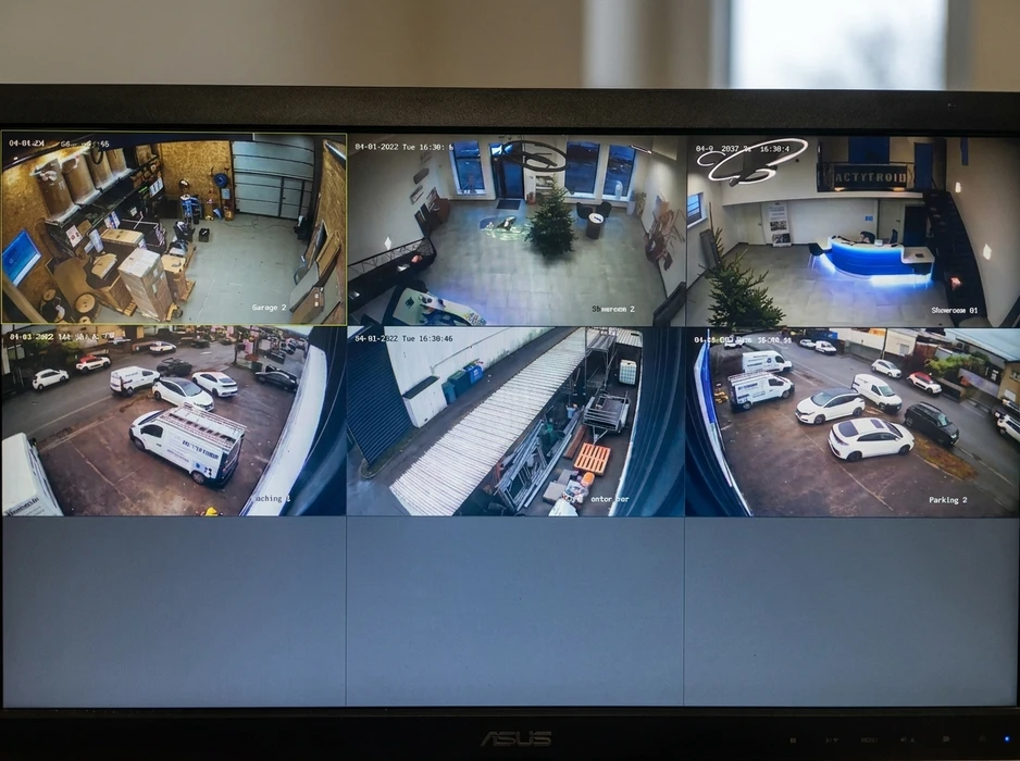 Écran de supervision affichant 6 flux CCTV simultanés : garage de stockage, hall d'accueil avec sapin, showroom avec comptoir lumineux bleu, parking extérieur avec véhicules utilitaires blancs, quai de chargement avec palettes, zone logistique